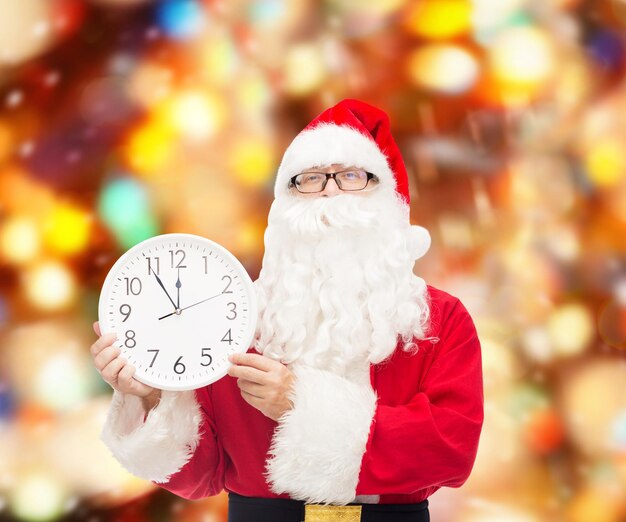 christmas, holidays and people concept - man in costume of santa claus with clock showing twelve over red lights background