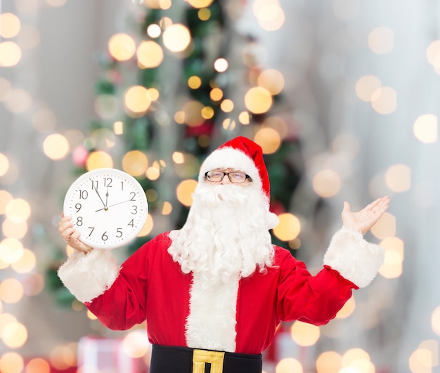 Concetto di natale, vacanze e persone - uomo in costume di babbo natale con orologio che mostra dodici su sfondo di luci ad albero