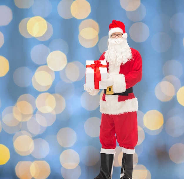 christmas, holidays and people concept - man in costume of santa claus with gift box over blue lights background