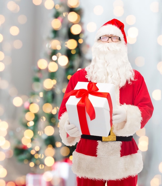 christmas, holidays and people concept - man in costume of santa claus with gift box over tree lights background