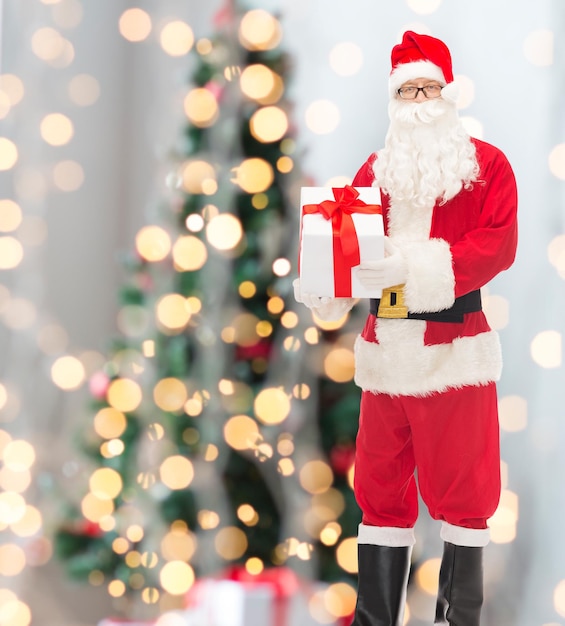 christmas, holidays and people concept - man in costume of santa claus with gift box over tree lights background