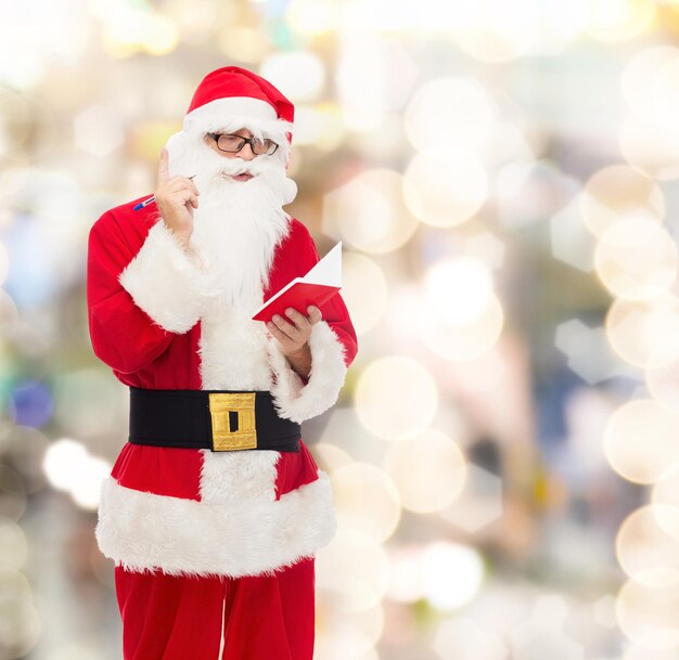 christmas, holidays and people concept - man in costume of santa claus with notepad and pen over lights background