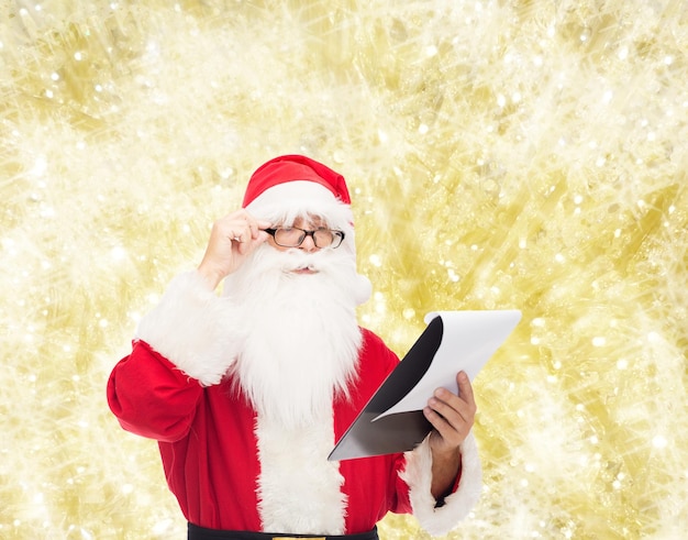 Christmas, holidays and people concept - man in costume of santa claus with notepad over yellow lights background