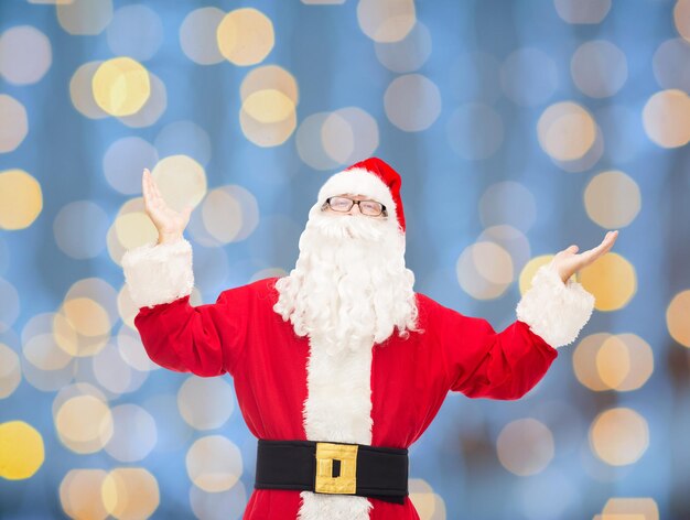 christmas, holidays and people concept - man in costume of santa claus with raised hands over blue lights background