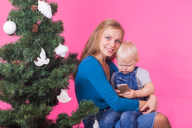 クリスマス、休日、技術、人々の概念-クリスマスツリーの近くで娘を抱き締める若い幸せな女性。モバイルを使用している子供。