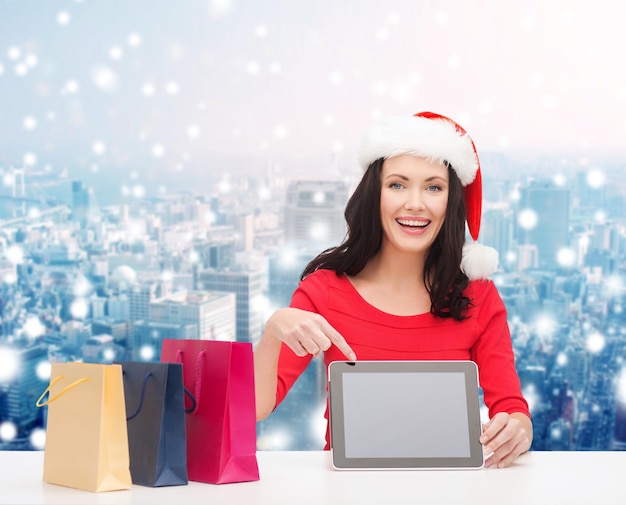 christmas, holidays, technology, advertising and people concept - smiling woman in santa helper hat with shopping bags and tablet pc computer over snowy city background