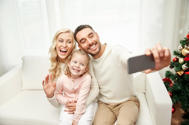 christmas, holidays, technology and people concept - happy family sitting on sofa and taking selfie picture with smartphone at home