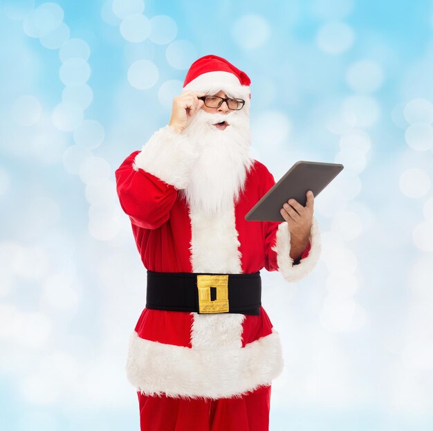 christmas, holidays, technology and people concept - man in costume of santa claus with tablet pc computer over blue lights background