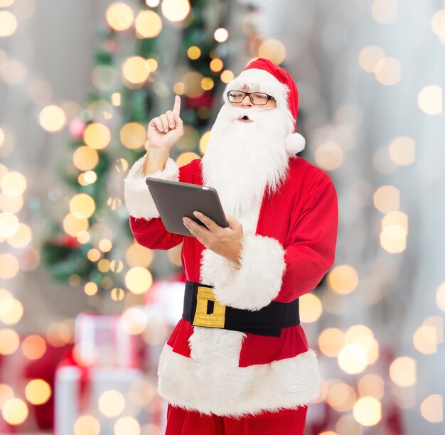 christmas, holidays, technology and people concept - man in costume of santa claus with tablet pc computer pointing finger up over tree lights background