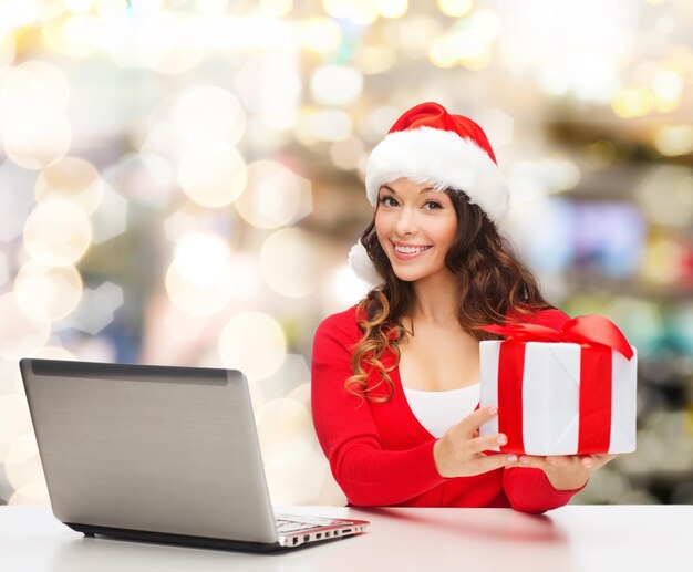 christmas, holidays, technology and people concept - smiling woman in santa helper hat with gift box and laptop computer over lights background