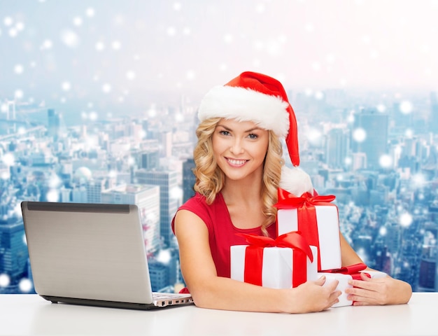 christmas, holidays, technology and people concept - smiling woman in santa helper hat with gifts and laptop computer over snowy city background