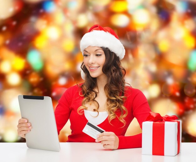 christmas, holidays, technology and shopping concept - smiling woman in santa helper hat with gift box, credit card and tablet pc computer over red lights background