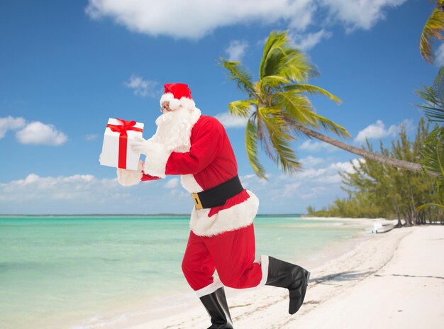 christmas, holidays, travel and people concept - man in costume of santa claus running with gift box over tropical beach background