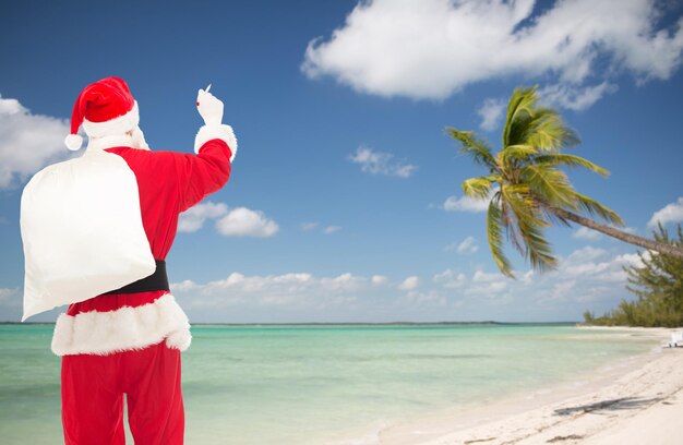 Photo christmas, holidays, travel and people concept - man in costume of santa claus with bag writing something from back over tropical beach background