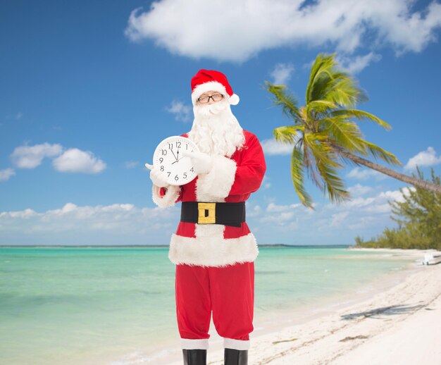 christmas, holidays, travel and people concept - man in costume of santa claus with clock showing twelve pointing finger over tropical beach background