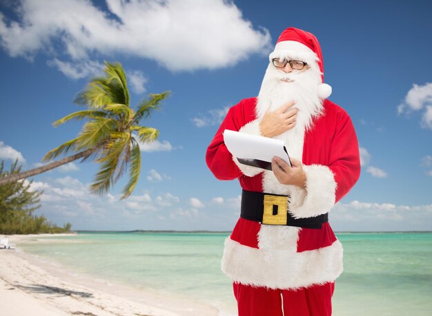 christmas, holidays, travel and people concept - man in costume of santa claus with notepad over tropical beach background