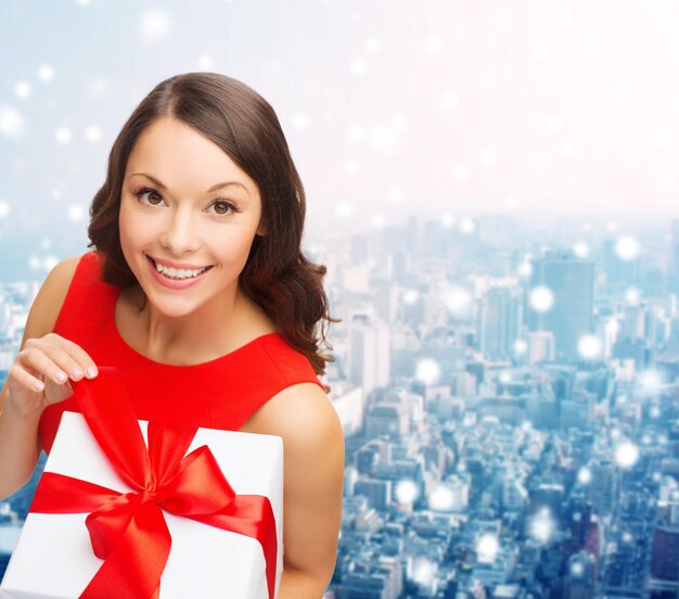 christmas, holidays, valentine's day, celebration and people concept - smiling woman in red dress with gift box over snowy city background