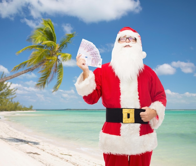 christmas, holidays, winning, currency and people concept - man in costume of santa claus with euro money over tropical beach background