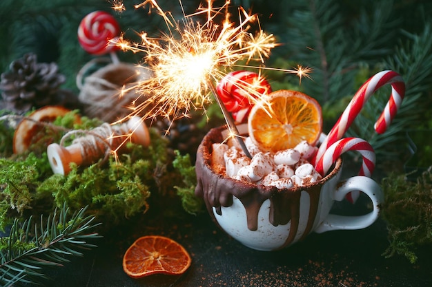 Christmas hot chocolate in a white cup with marshmallow, caramelized oranges and candy cane with sparklers on dark background, Selective focus