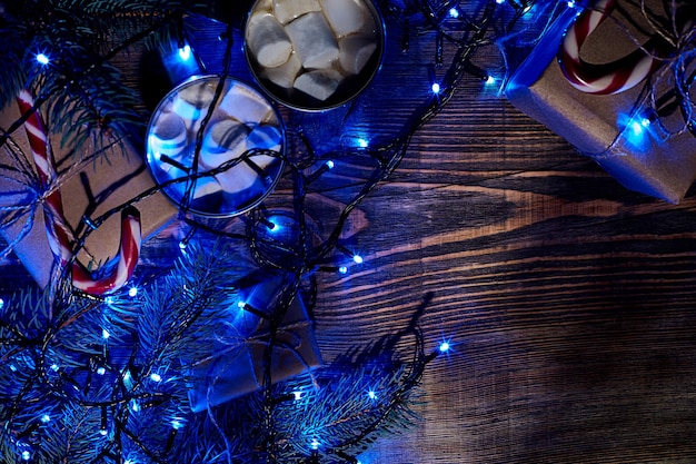 Christmas hot drink. Cocoa with marshmallow, gift box, christmas lights, garland and spruce branches on a wooden background. Top view. Copy space. Flat lay. Still life. Mock-up