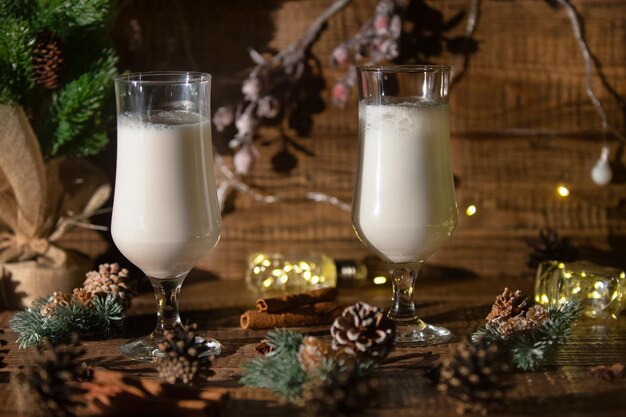 Christmas hot drink eggnog with cinnamon is poured into two glasses on a wooden background with spru