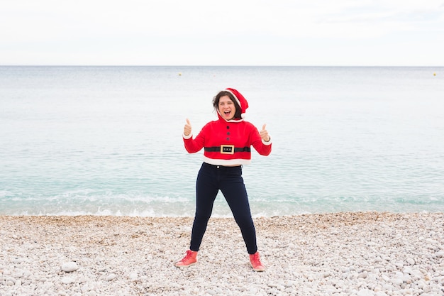 Concetto di natale, umorismo e persone - giovane donna felice in tuta di babbo natale in spiaggia che mostra i pollici in su vicino al mare.