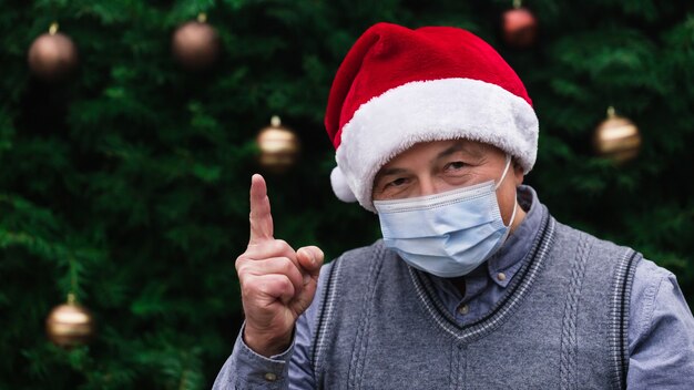 Christmas idea.Close up Portrait of senior man wearing a santa claus hat and medical mask with emotion. Against the background of a Christmas tree. Coronavirus pandemic