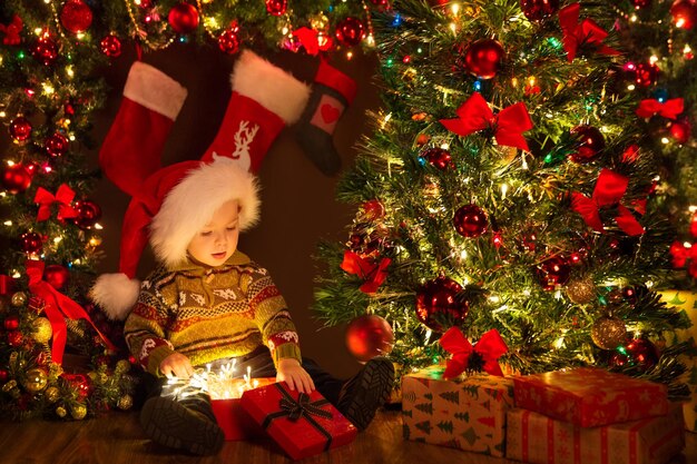 Christmas Kid opening Gift Box with Xmas Lights Garland. Cute Baby in Santa Hat sitting next to Decorated Fir Tree in Dark Room Interior with Shining Ornaments