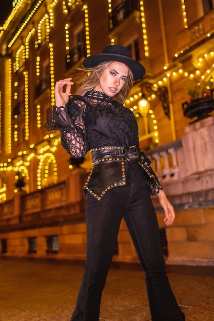 Christmas lights in the city, blonde girl dressed in black with a hat next to an illuminated building, winter lifestyle