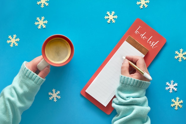 Christmas do list and woman's hands in mint color sweater with cup of coffee on blue table