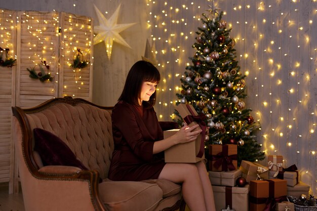 Christmas and magic concept - portrait of young woman opening gift box in decorated room with Christmas tree and led lights