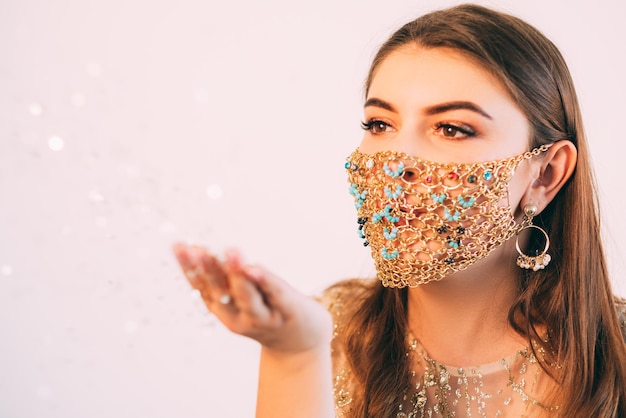 Christmas magic. Pandemic winter holidays. Make a wish. Beautiful woman in glamour gold chain face mask blowing bokeh light spell sparks from open palm on pastel pink copy space background.