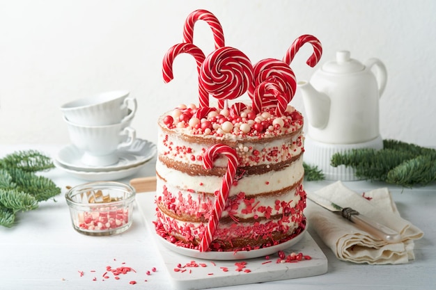 Christmas many layers cake decorated with lolli pops red and white confetti on white cake stand Christmas balls fir branches on white concrete table Selective focus