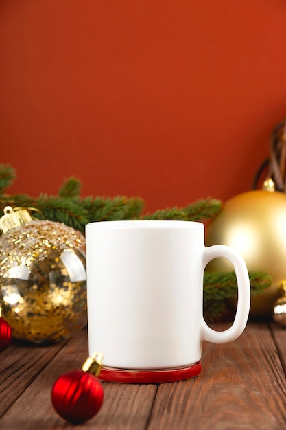 お祝いの装飾が施された木製のテーブルに白いマグカップのクリスマスモックアップ。赤い壁に金色のクリスマスボール。