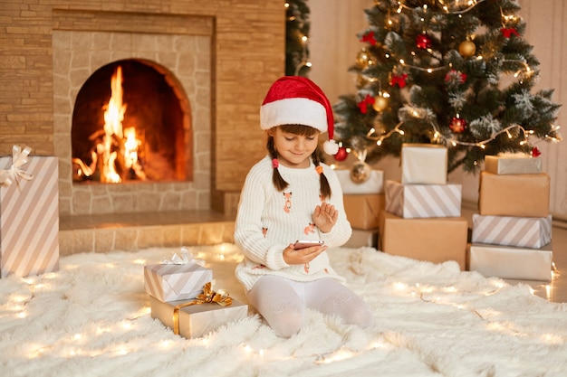 Atmosfera natalizia. piccolo bambino sveglio che tiene il telefono nelle mani, prendi uno smart phone come regalo di natale, seduto sul pavimento su un morbido tappeto vicino a scatole regalo e camino, veste il cappello di babbo natale festivo.