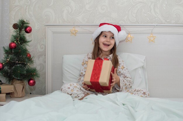 크리스마스 아침, 크리스마스 트리 배경에 침대에 선물을 들고 잠옷을 입은 어린 소녀. 행복하고 웃는 아이가 집에서 새해 선물을 엽니다. 휴일, 새해, 크리스마스의 개념