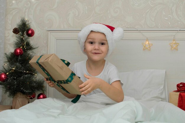 Christmas morning, little girl in pajamas with gifts in bed against the background of Christmas tree. A happy, smiling child opens New Year's gift at home. The concept of holidays, New Year, Christmas