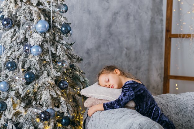 Christmas morning. Tired little girl sleeping on couch, waiting for Santa with gifts at decorated fir tree at home.