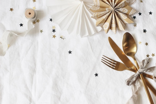 Christmas New Year composition Decorative table setting Paper stars golden cutlery and confetti stars on white linen background Celebration birthday party concept Flat lay top view copy space