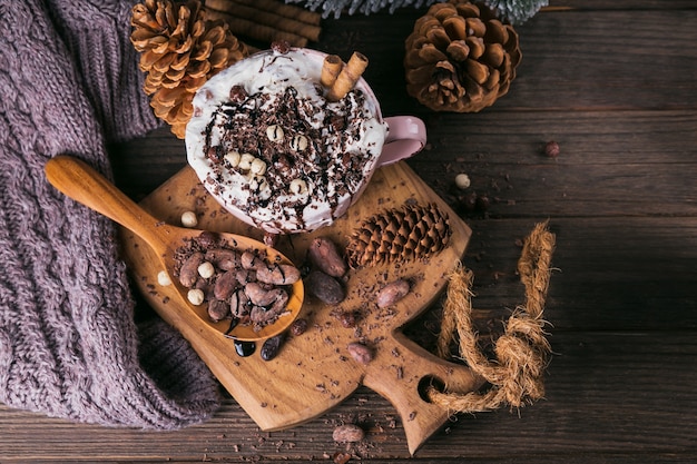 Christmas or New Year composition with hot chocolate or cocoa drink with whipped cream served with chopped chocolate and cacao beans on rustic wooden plate.  Top view