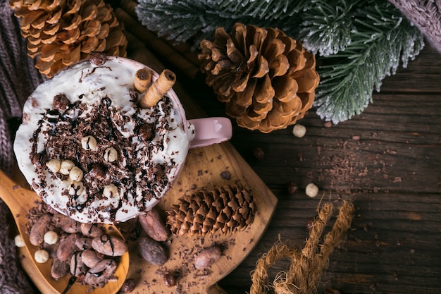 Christmas or New Year composition with hot chocolate or cocoa drink with whipped cream served with chopped chocolate and cacao beans on rustic wooden plate. 