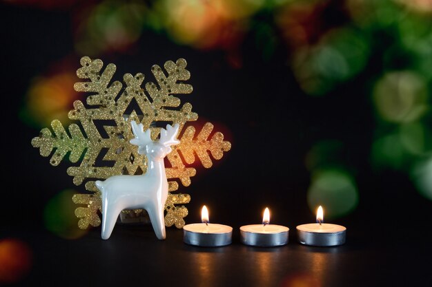 Christmas, New Year composition with three candles, white porcelain reindeer and golden snowflake on black background.