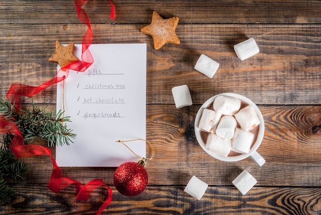 Christmas, New Year concept. Wooden table, notebook with to do list  gingerbread, gifts, hot chocolate, Christmas tree , cocoa mug, xmas ball, pine tree, red ribbon, marshmallow. Top view  copyspace