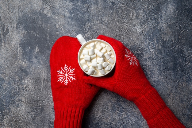 Christmas and New Year cozy holiday composition with scarf, woman hands in mittens, mugs with hot drink and marshmallow on the gray concrete background. Flat lay, top view.