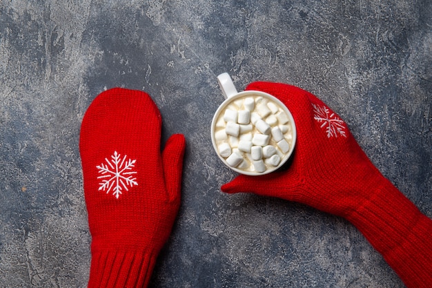 Christmas and New Year cozy holiday composition with scarf, woman hands in mittens, mugs with hot drink and marshmallow on the gray concrete background. Flat lay, top view.