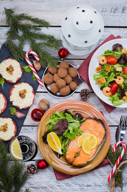Concetto della tabella della regolazione della cena della famiglia del nuovo anno o di natale con la decorazione di festa. salmone, insalata, aperitivi e dessert deliziosi dell'arrosto di bistecca su di legno bianco. vista dall'alto
