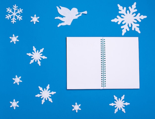 Photo christmas and new year flat composition with white blank notepad, white angel playing trumpet and christmas snowflakes on blue background