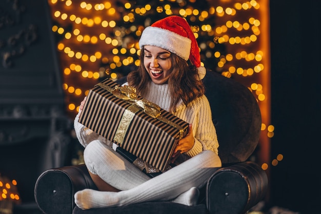 Christmas, New Year. Pretty woman in warm sweater sitting on the chair at home with gift box