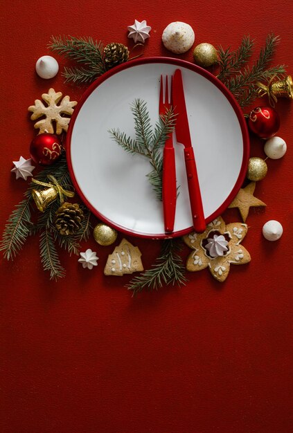 Foto tavola apparecchiata per natale e capodanno. piatto vuoto bianco con posate e rami di albero di natale, tovagliolo rosso su fondo di cemento rosso.