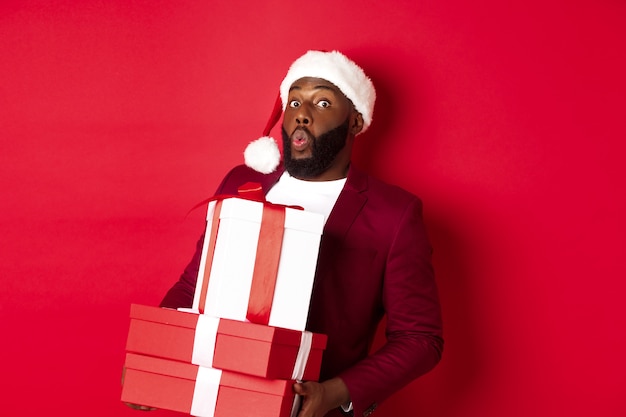 Natale, capodanno e concetto di shopping. felice uomo di colore con cappello da babbo natale e blazer che tiene regali di natale, porta regali e sorride, in piedi su sfondo rosso